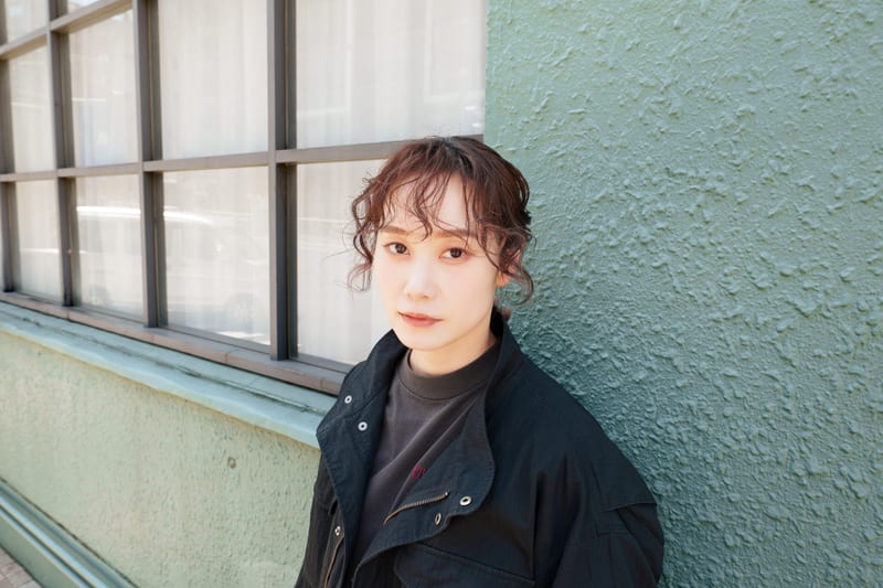 A person in a black jacket leans against a pale green wall beside a row of windows