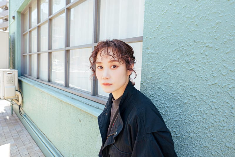 Against a pale green wall and windows, a person in a black jacket looks into the camera