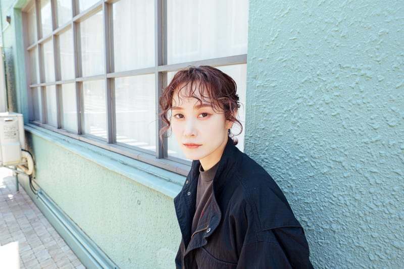 A person in a black jacket stands by a pale green wall, sunlight on the pavement