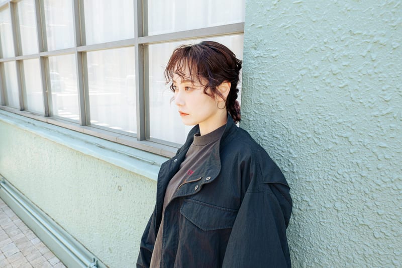Eyes lowered, a person in a black jacket leans on a pale green wall by a window
