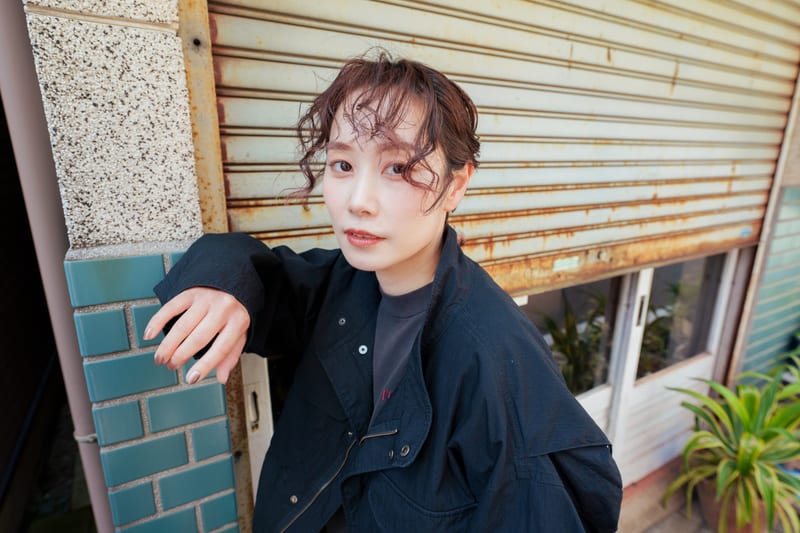 In front of a rusted metal shutter, a person rests a hand on a green tiled pillar