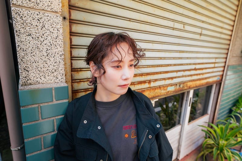 In front of a rusted shutter, a person in a black jacket looks down to the side