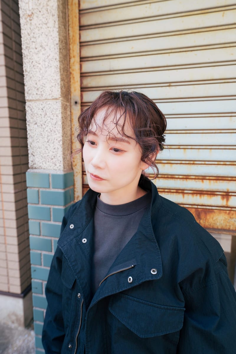 Head turned aside, a person in a black jacket stands by a rusted metal shutter