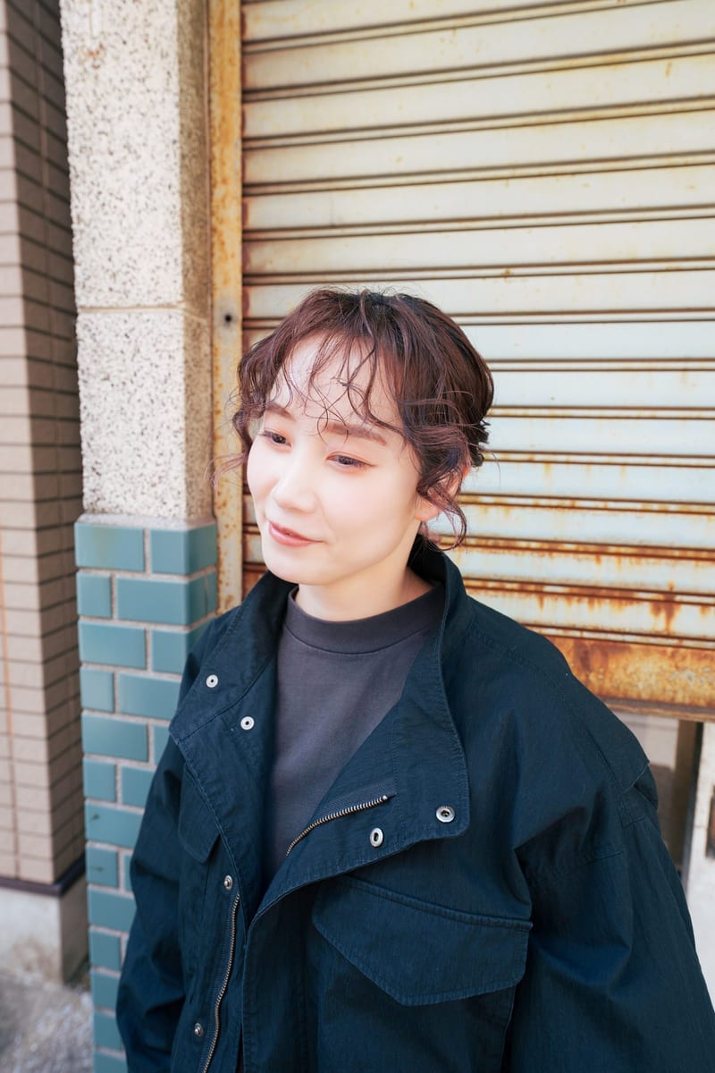 Smiling to the side, a person in a black jacket in front of a rusted shutter