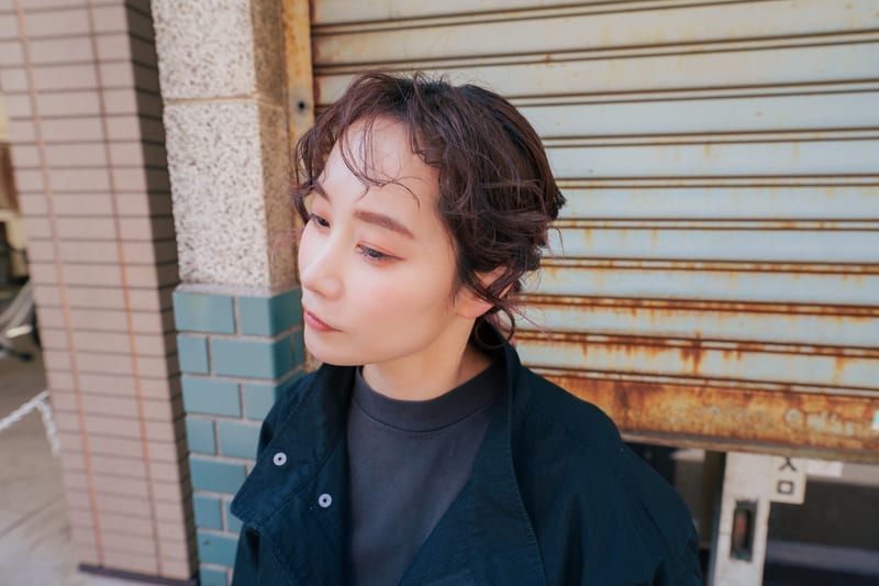Close up in profile, a person with curly brown hair in front of a rusted shutter
