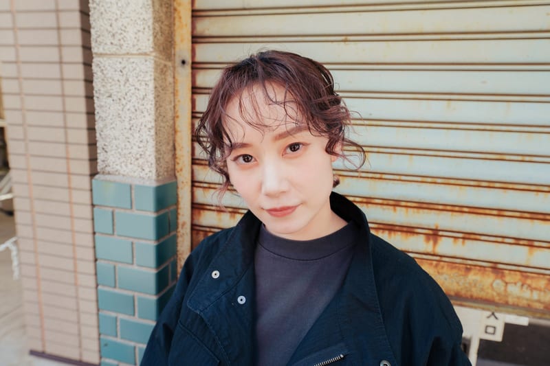 Close up, a person with curly brown hair looks into the camera in front of a rusted shutter