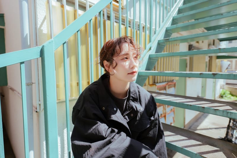 Sitting on a green outdoor staircase in bright sun, a person looks off to the side