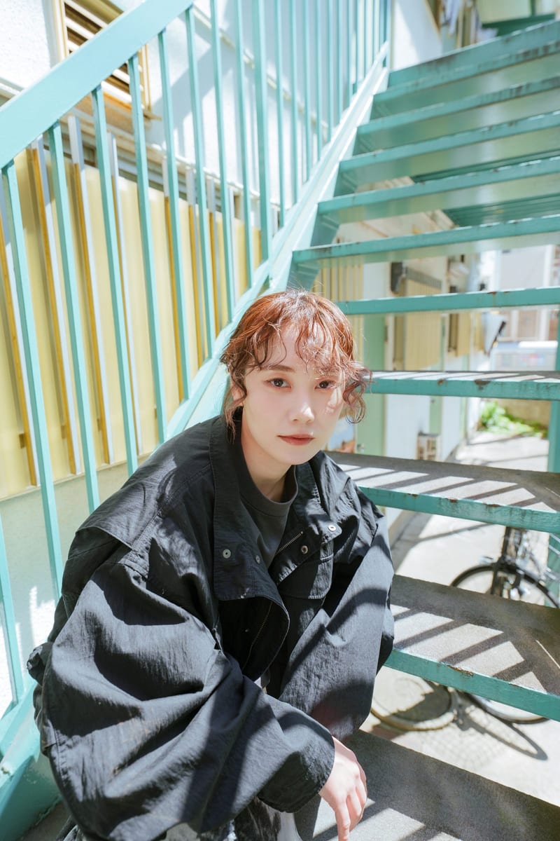 Crouched on green metal stairs, a person in a black jacket looks into the camera