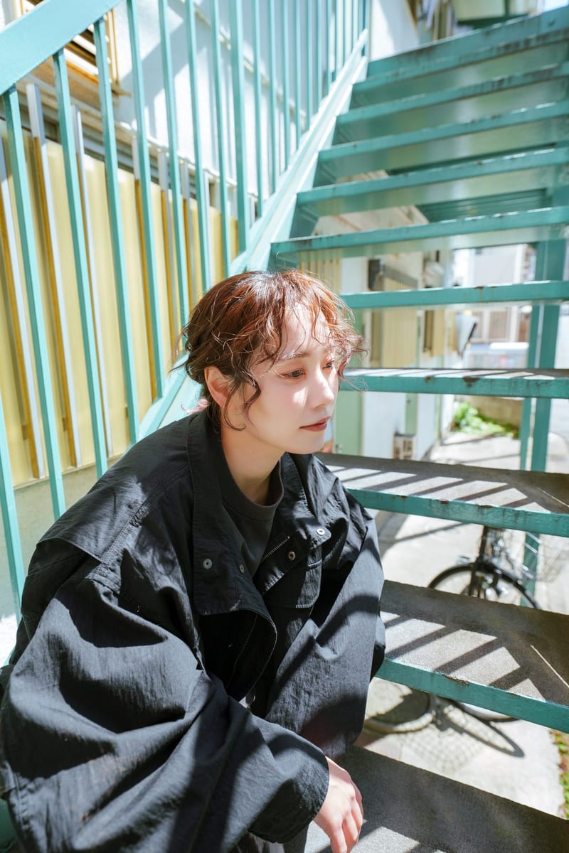 Crouched on green metal stairs in sunlight, a person looks down to the side