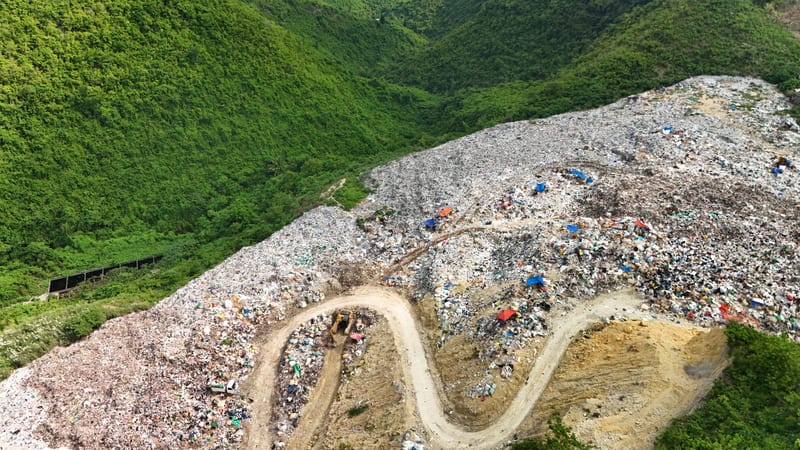 From the air, a landfill spreads down a green valley with a dirt road winding through it