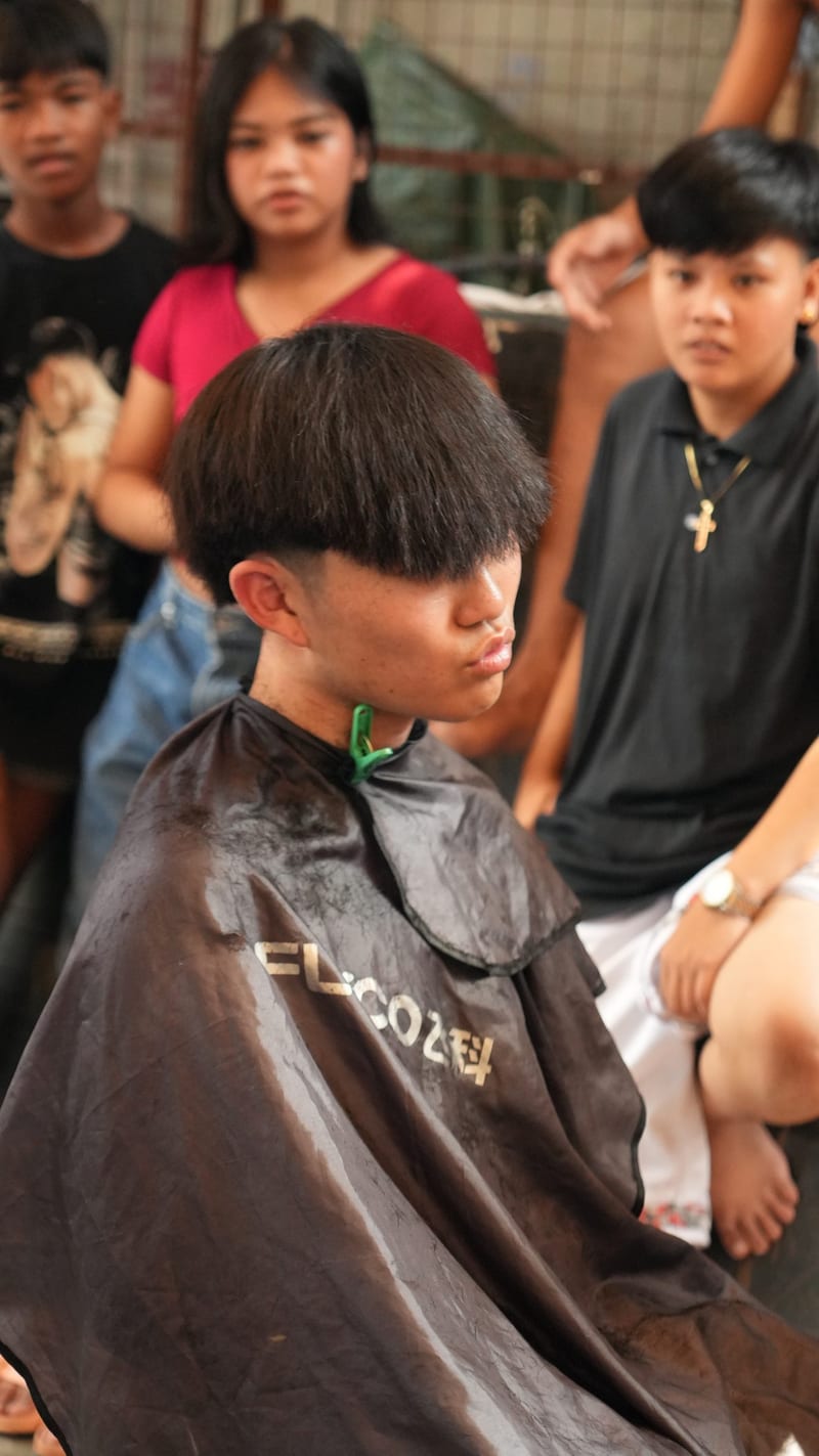A boy in a barber's cape has his hair cut on a Manila street while friends look on