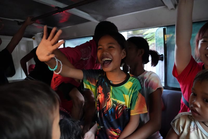 Inside a crowded jeepney, a girl in a bright shirt laughs and waves at the camera