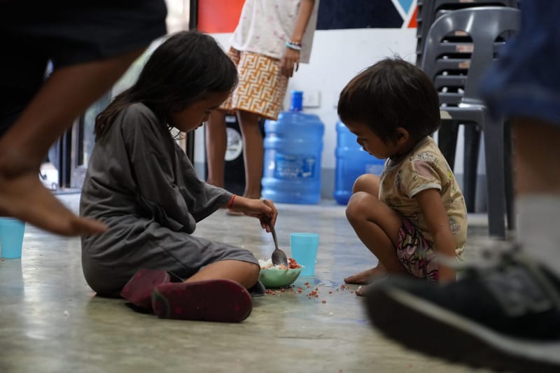 Two small children squat on a concrete floor with plastic cups, a water jug behind them