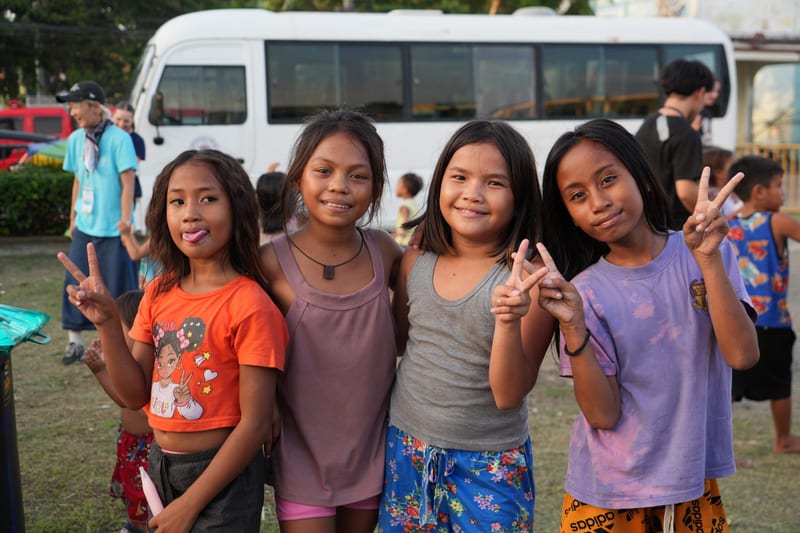 Four girls in orange, grey and lilac make peace signs in front of a white bus