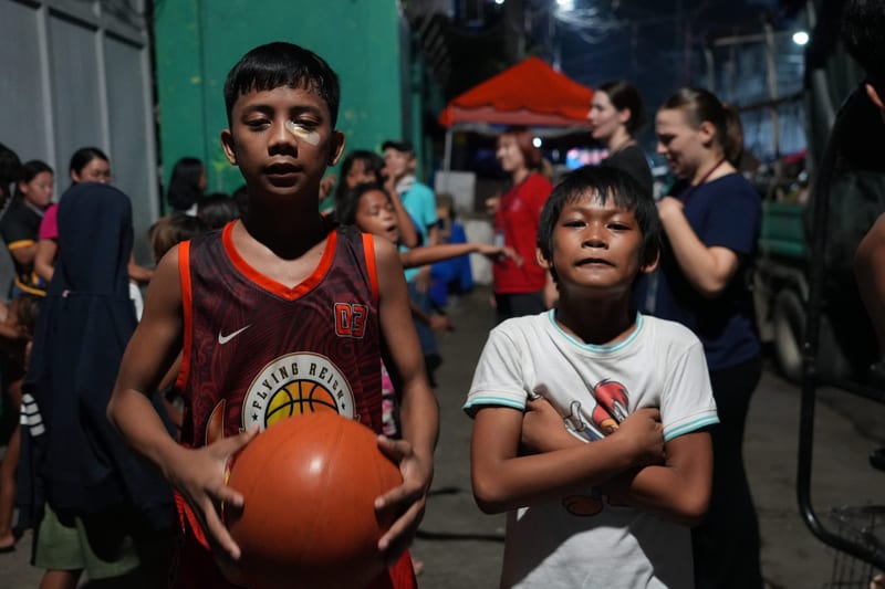 At night on a street, a boy holds a basketball while a smaller boy beside him folds his arms