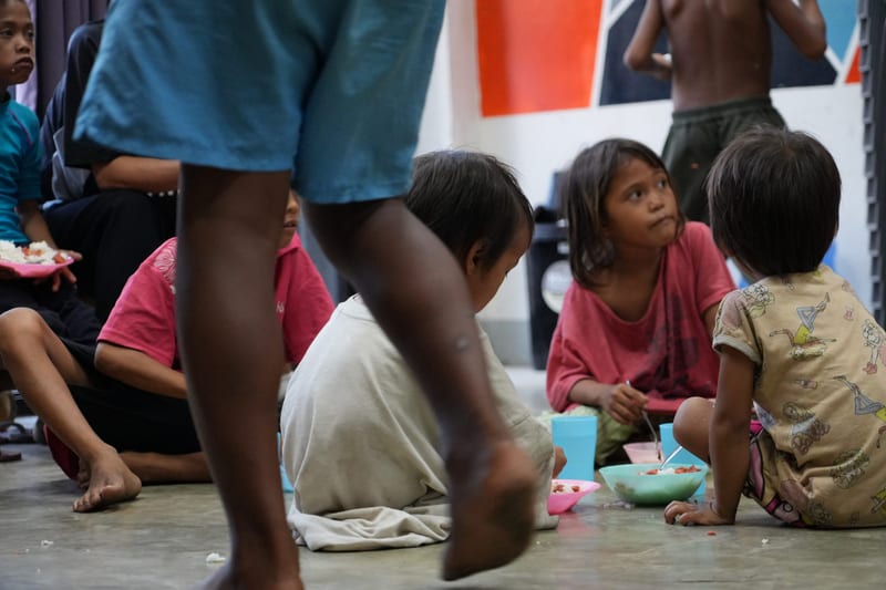 Children sit on the floor with plates of food while adults walk past