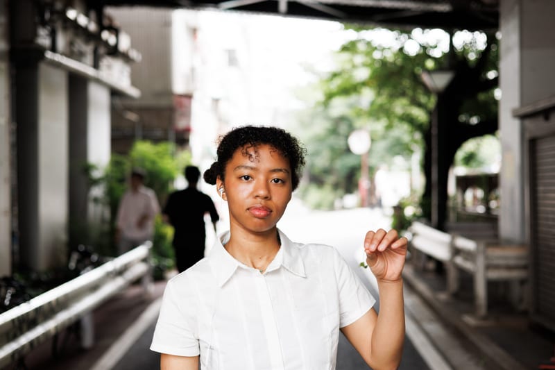 A person in a white shirt stands under a railway bridge on a quiet street, one hand raised