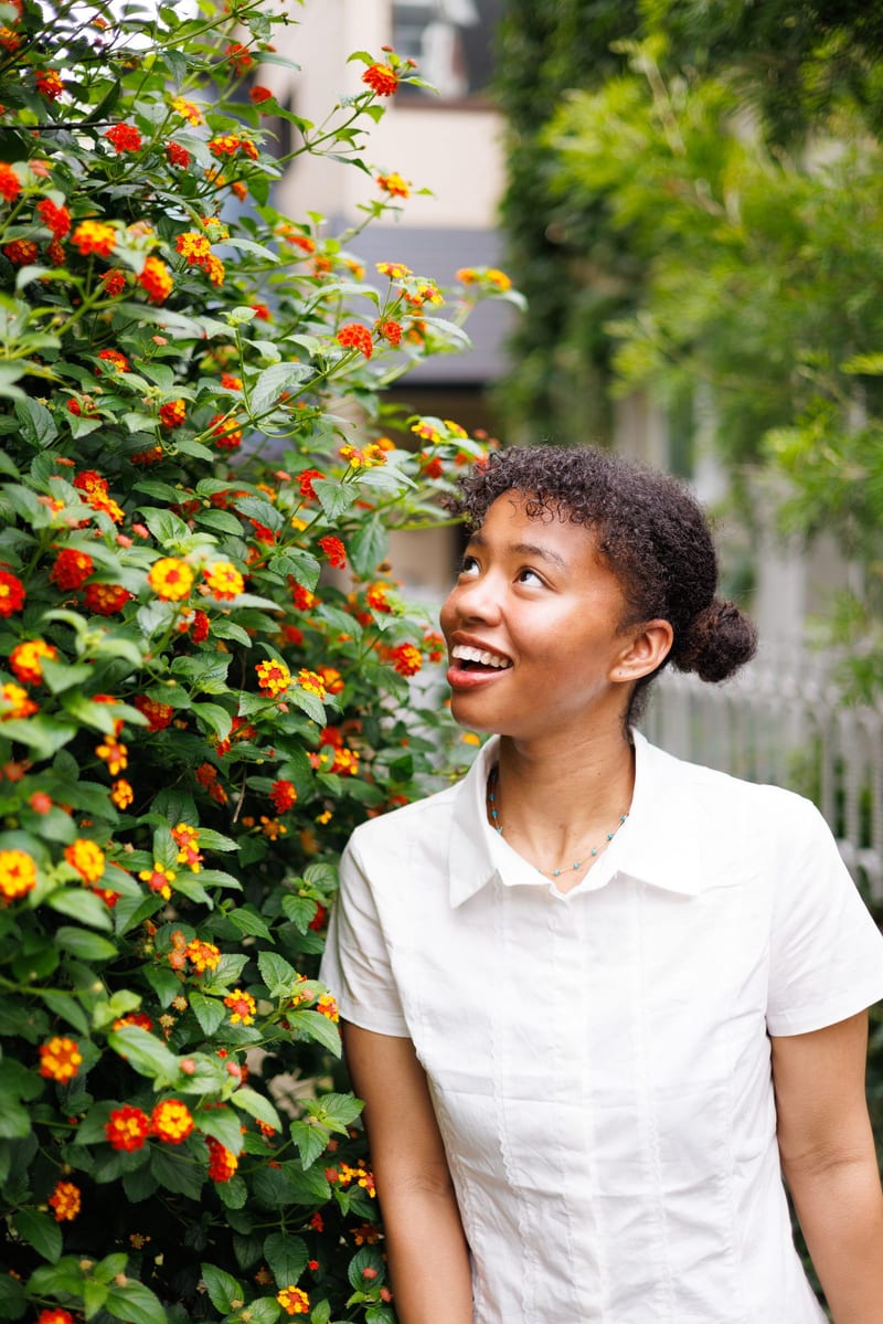 Beside a hedge of small orange flowers, a person in a white shirt looks up and laughs