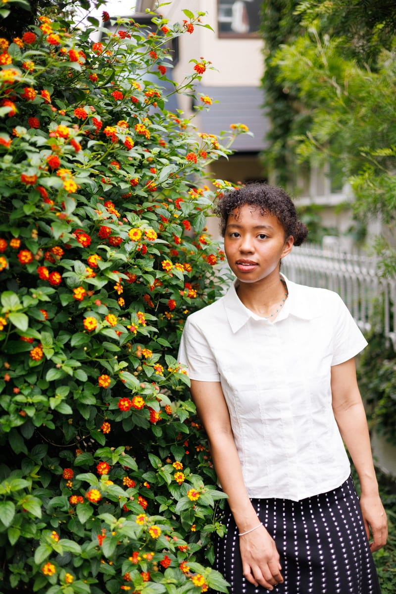 A person in a white shirt and a dotted skirt stands beside a flowering hedge