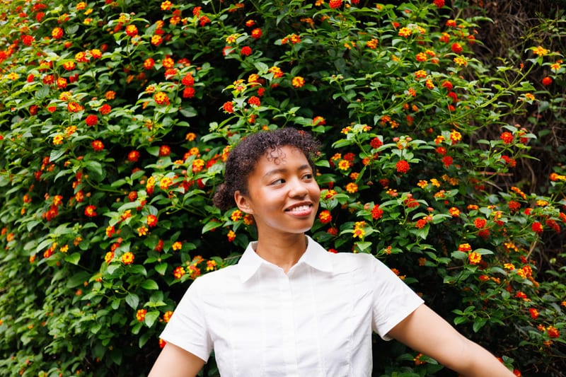 A person in a white shirt spreads their arms in front of a hedge covered in small orange flowers