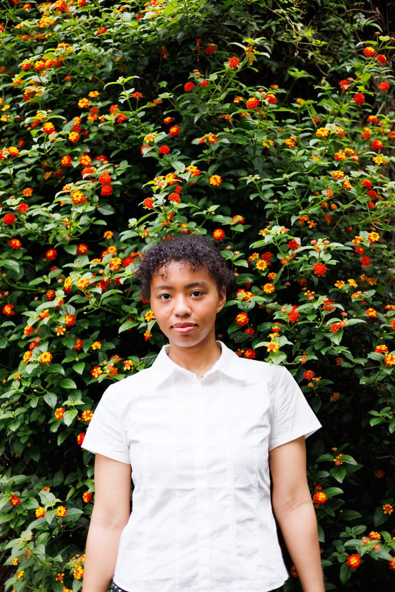 Facing the camera, a person in a white shirt stands in front of a hedge of orange flowers