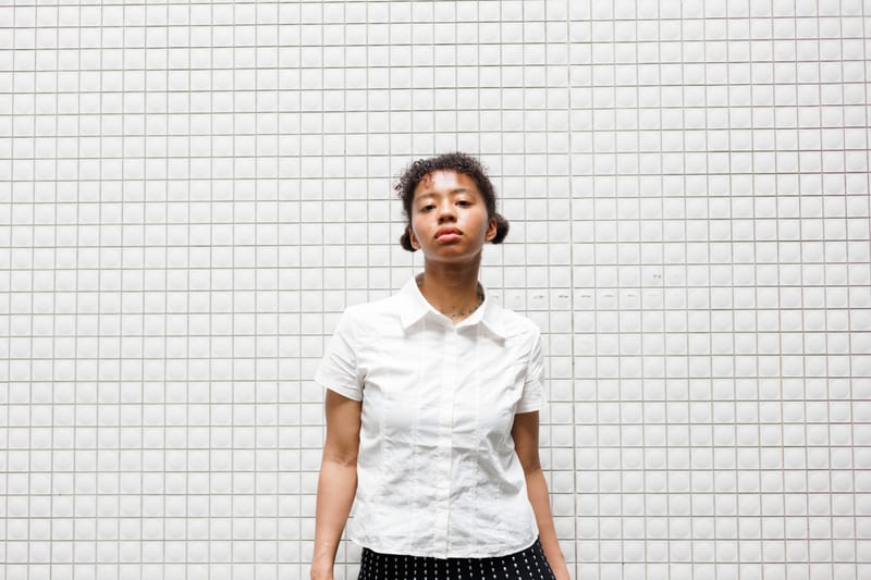 A person in a white shirt stands straight against a white tiled wall