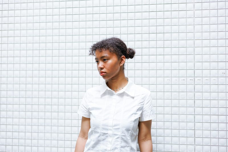 Against a white tiled wall, a person in a white shirt looks to the side
