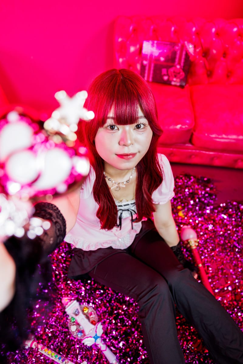 A heart wand blurred in the foreground, a person with red hair smiles on the floor of a hot pink room