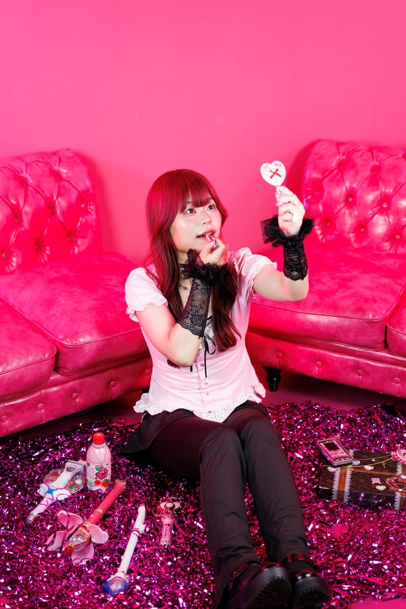 Sitting on pink tinsel between two pink sofas, a person holds a small heart toy up beside their face