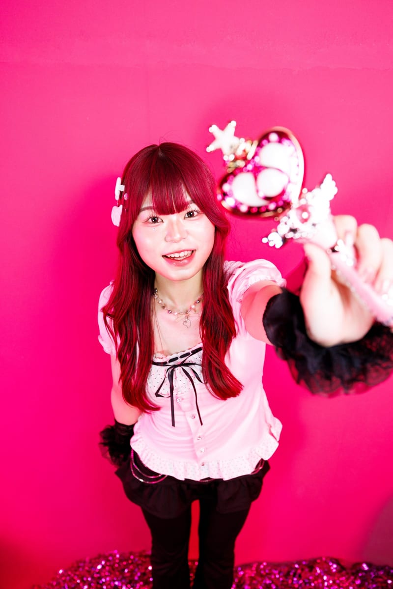 Standing against a hot pink wall, a person reaches a heart wand out to the camera