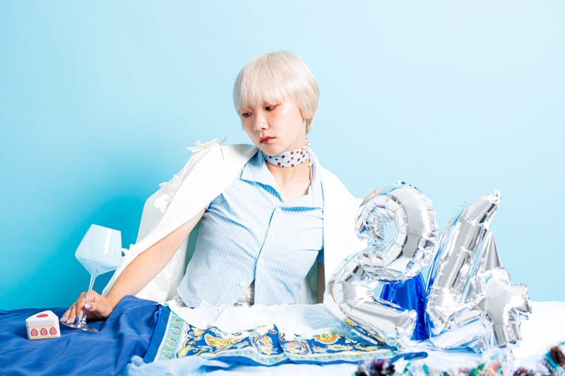 Seated at a table draped in blue cloth, a person reaches for a glass beside silver number balloons