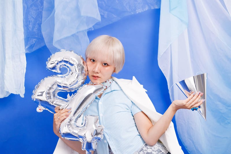 In front of a blue backdrop and sheer curtains, a person holds silver balloons and a silver cone