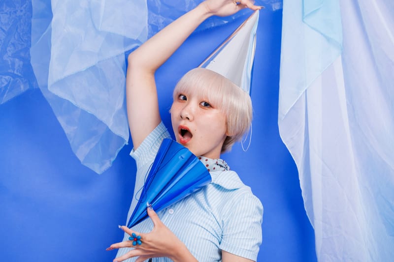 One arm raised with a silver cone hat, mouth open, a person poses against a blue backdrop