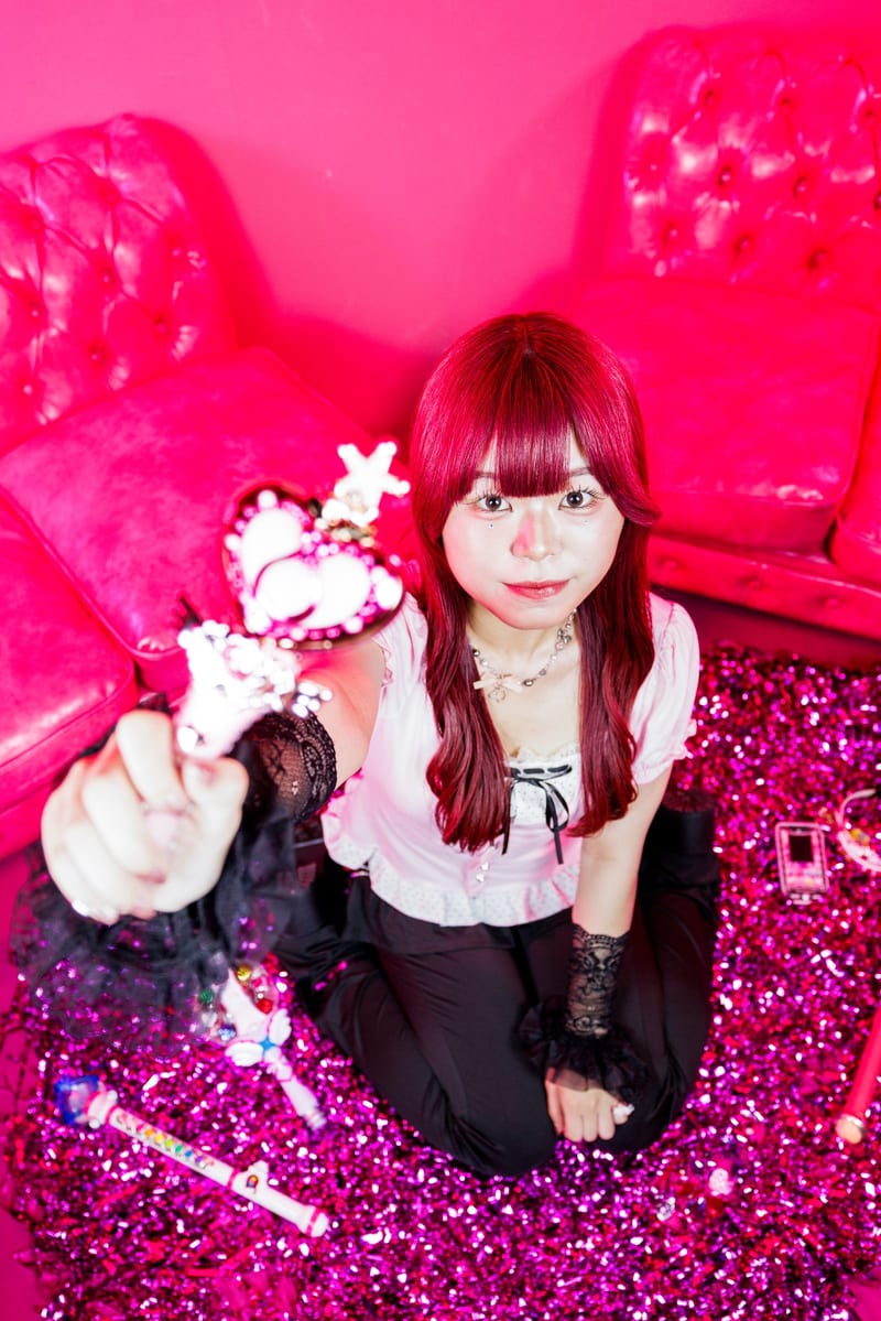Kneeling on pink tinsel in a hot pink room, a person with red hair holds a heart-shaped toy wand toward the camera