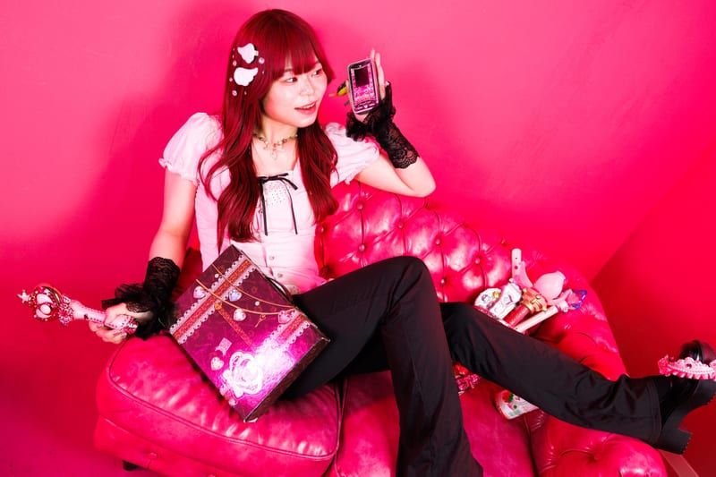 Sprawled on a pink sofa with a gift box on their lap, a person holds a pink flip phone beside their face