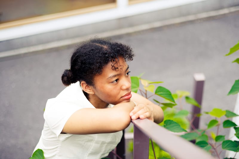 Leaning on a railing among green leaves, a person rests their chin on their arms