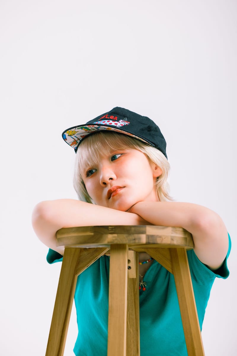 Arms folded on top of a wooden stool, a person in a black cap rests their chin and looks away