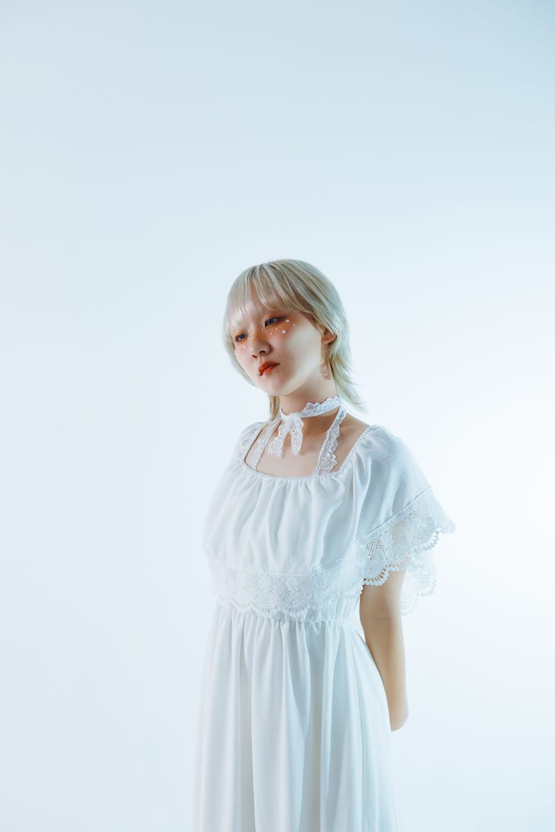A person in a white dress stands alone against a pale backdrop