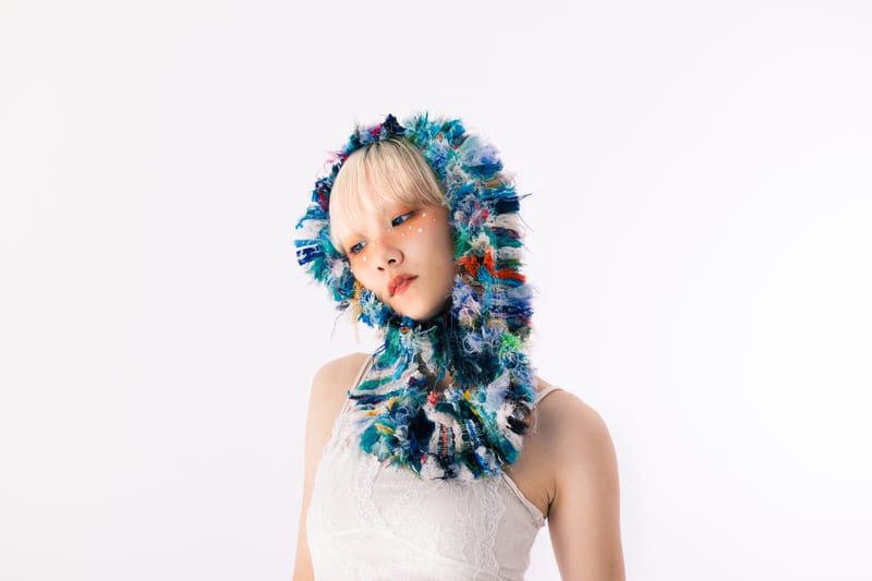 In a hood knitted from blue, white and green yarn, a person tilts their head against a white background