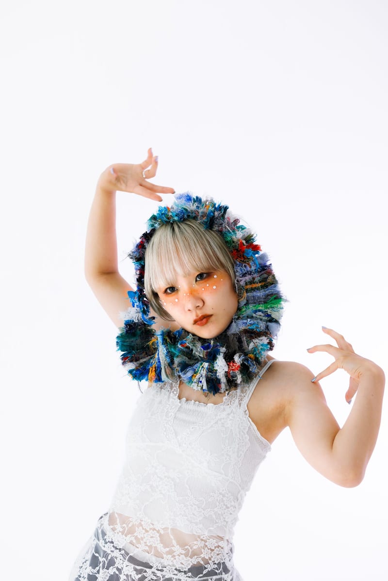 In a knitted hood, a person poses with both hands raised against a white background