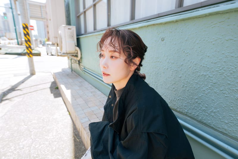 Sitting by a pale green wall on a sunny street, a person in a black jacket looks to the side