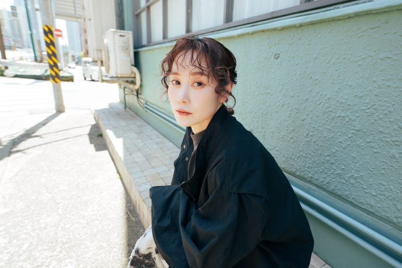 A person in black sits against a pale green wall in a sunlit back alley