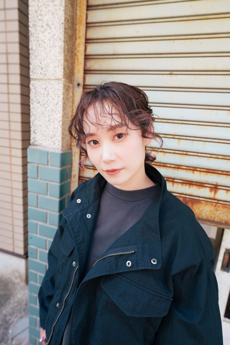 A person in a black jacket tilts their head toward the camera in front of a rusted shutter