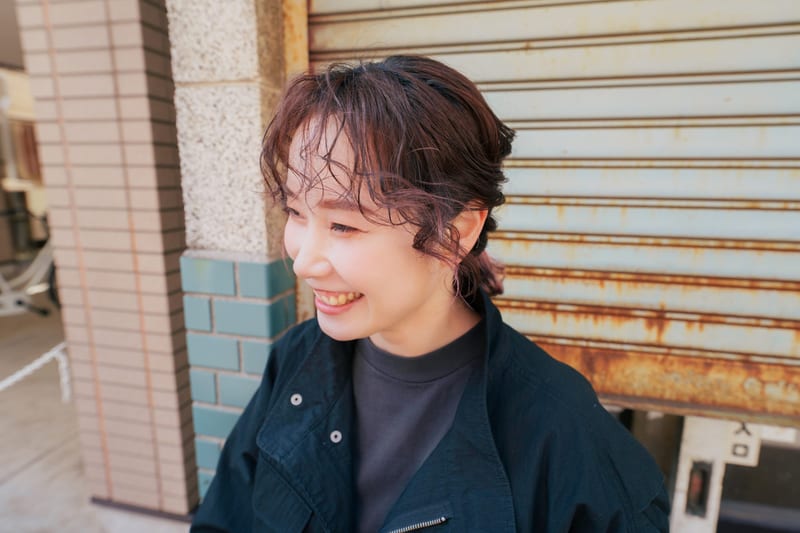 Laughing and looking away, a person in a black jacket in front of a rusted shutter