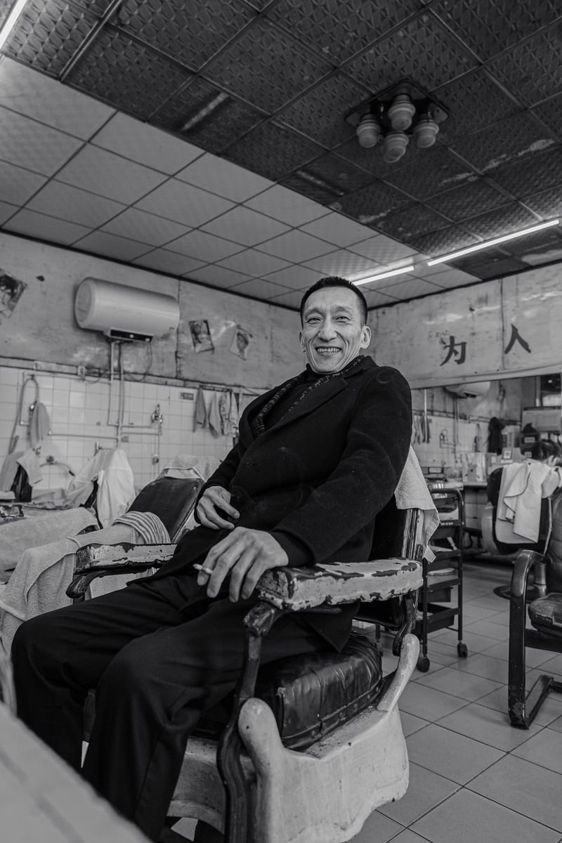 Black and white: an older man in a dark coat sits in a barber's chair and smiles