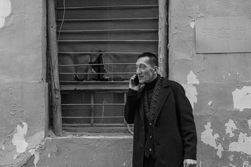 Black and white: a man in a dark coat talks on the phone in front of a barred window and a peeling wall