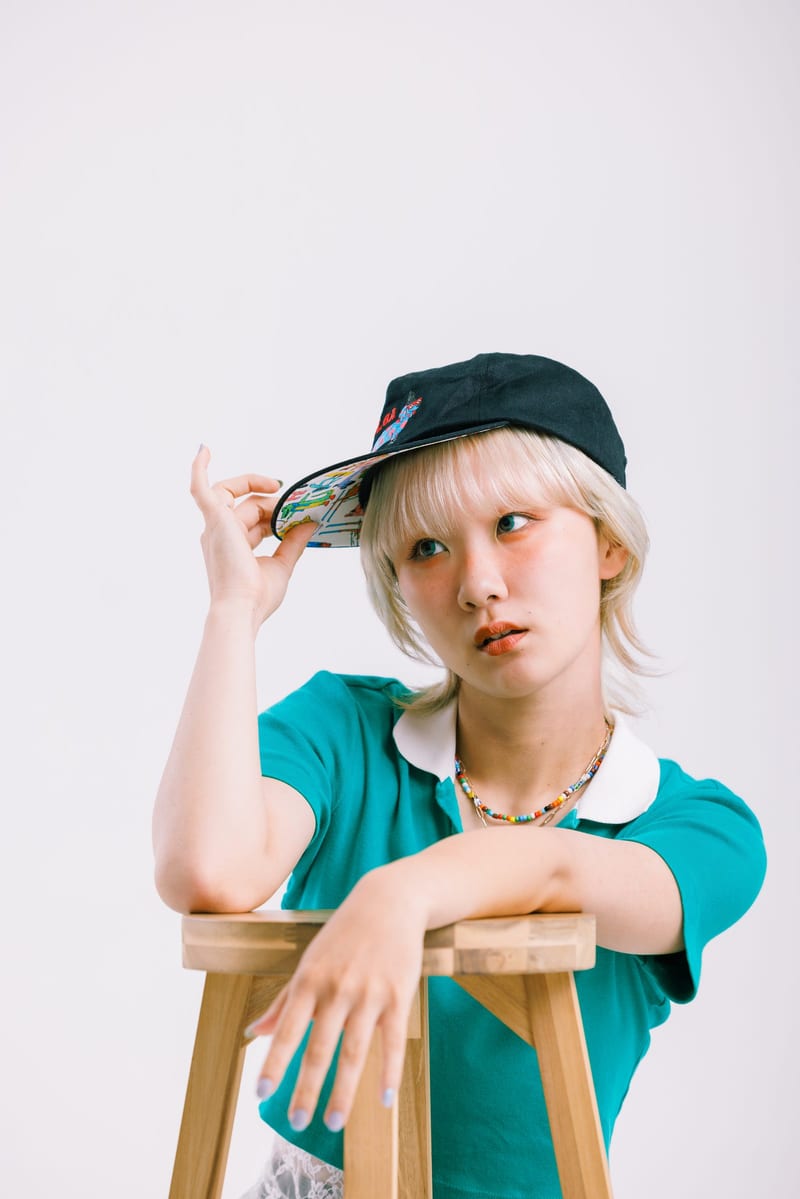 Leaning on a wooden stool, a person in a black cap touches its brim and glances aside