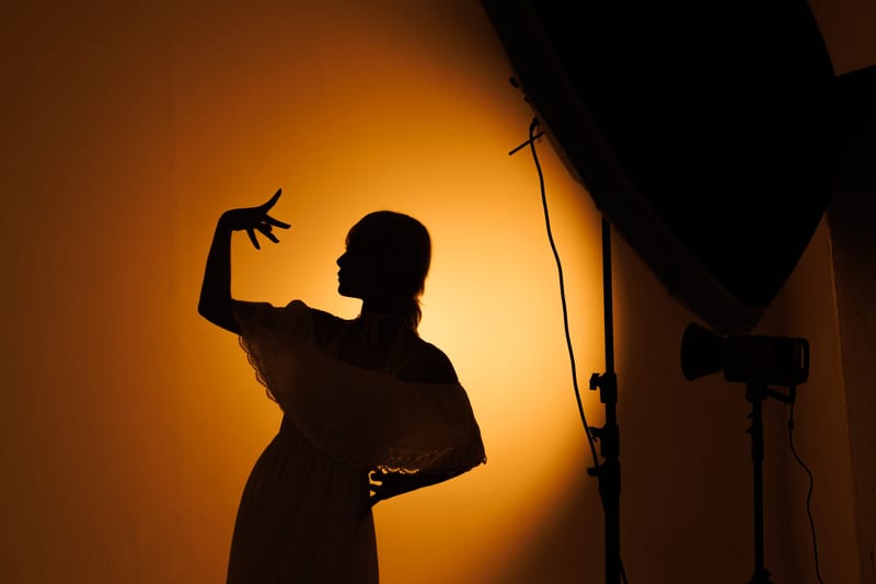 The silhouette of a person in a dress, one hand raised, against an orange spotlight in a studio