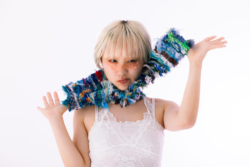 One arm raised, a person lifts the end of a long knitted scarf beside their face