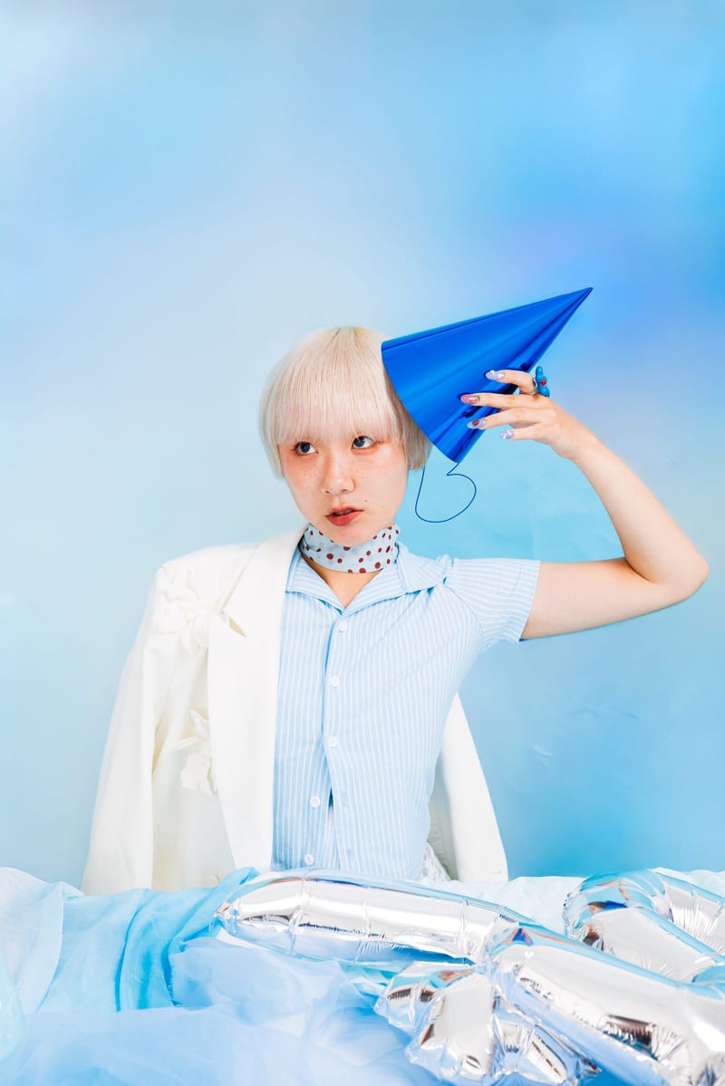 A person with pale blond hair holds a blue paper cone to their head against a pale blue backdrop, silver balloons at their feet